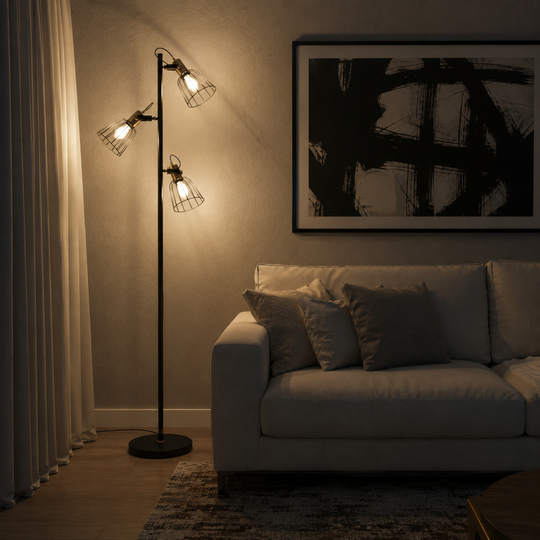 A modern black metal floor lamp with two adjustable arms and wire mesh cone shades, glowing with warm light in a dimly lit living room next to a white sofa and a framed black-and-white wall art piece.