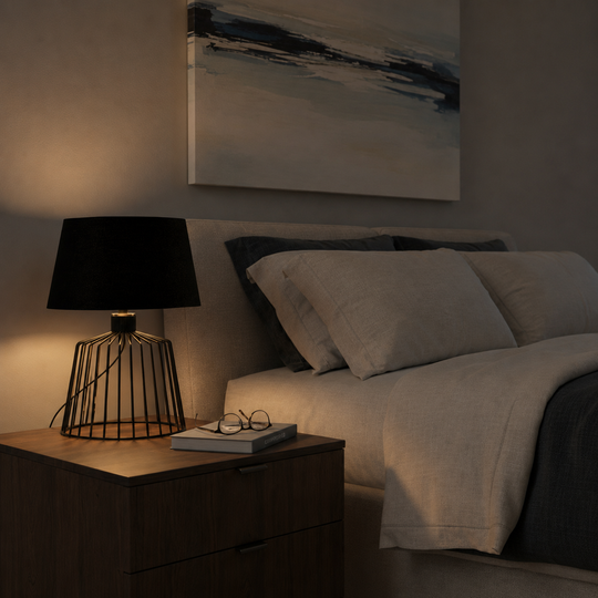 A modern black table lamp with a wire cage base sits on a wooden nightstand next to a bed with neutral bedding in a dimly lit bedroom.