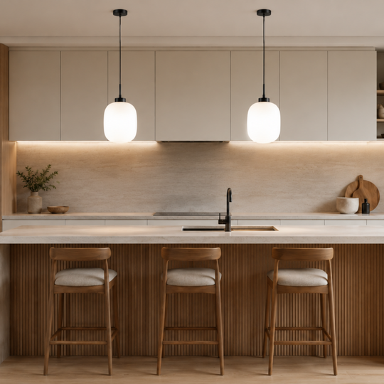 A modern kitchen interior with light wood cabinetry, a central island, three wooden bar stools with light cushions, pendant lights, and a large potted plant near a window.