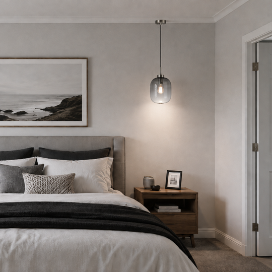 A styled bedroom interior featuring a neatly made bed with white and gray bedding, a gray upholstered headboard, two wooden nightstands, pendant lights, and a framed black-and-white coastal landscape print on the wall.