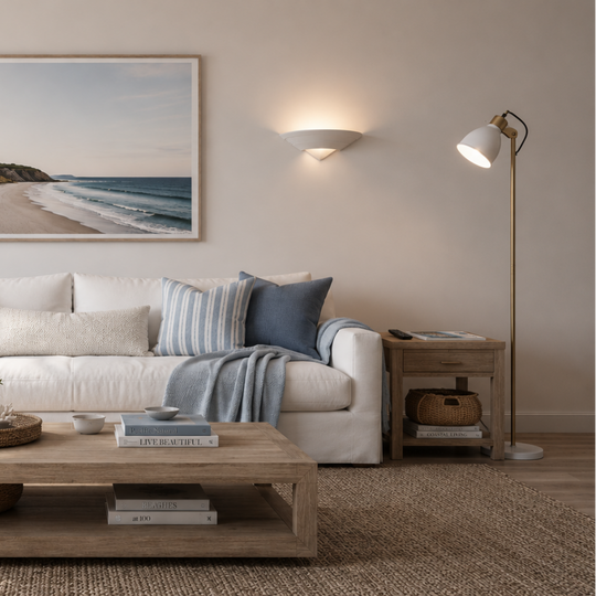 A coastal beach landscape print in a light wood frame hangs on a beige wall above a white sofa styled with blue and striped pillows in a bright, modern living room.