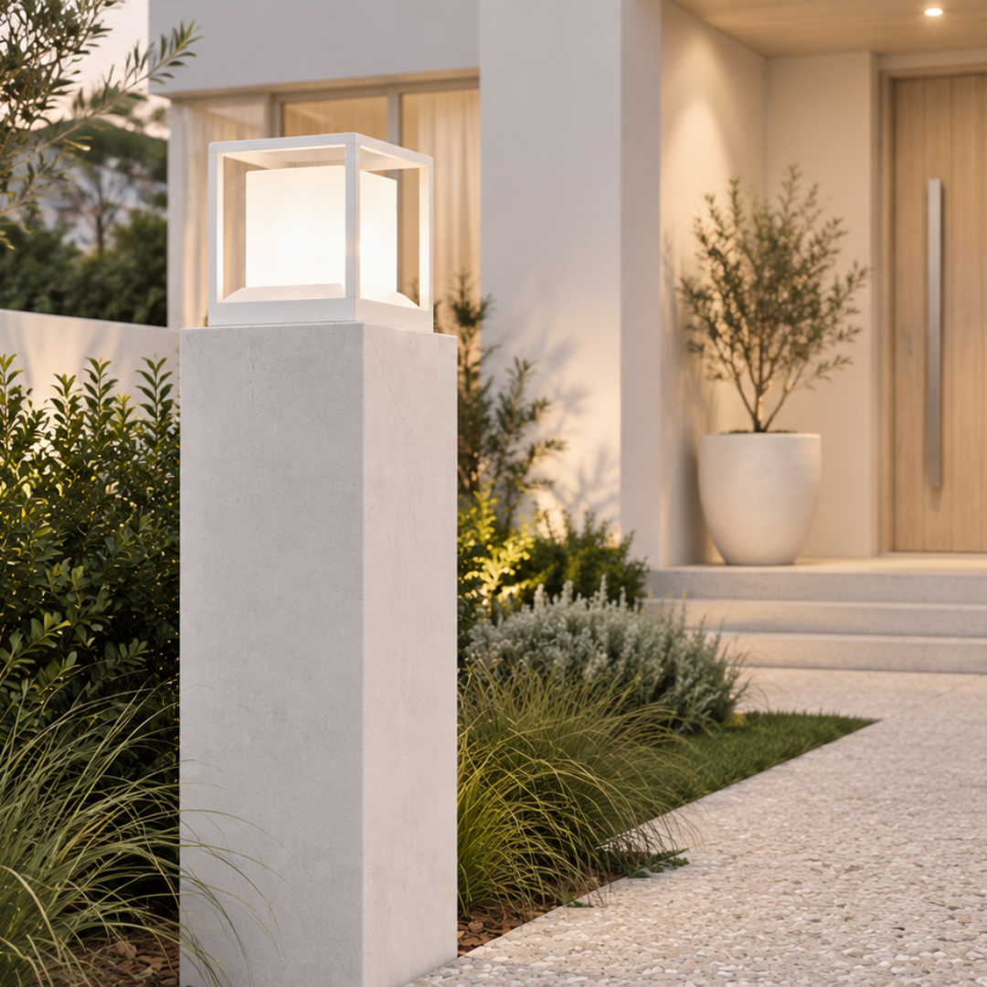 Modern outdoor architectural lighting featuring a square white pillar box lamp mounted on a white concrete post, set in a landscaped garden pathway leading to a house entrance.
