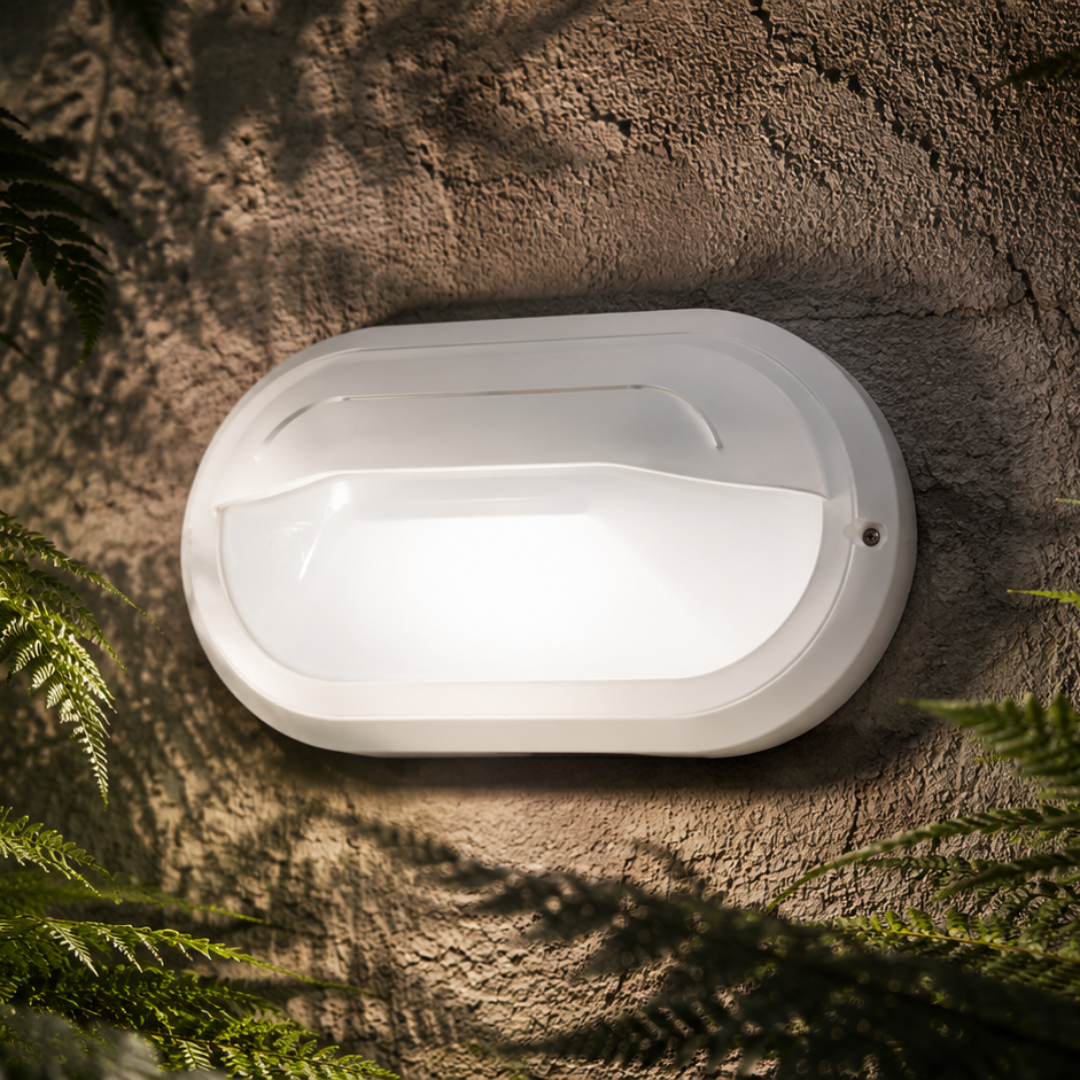 A white oval outdoor wall light fixture mounted on a textured stucco wall, surrounded by green fern leaves with dappled shadows cast across the scene.